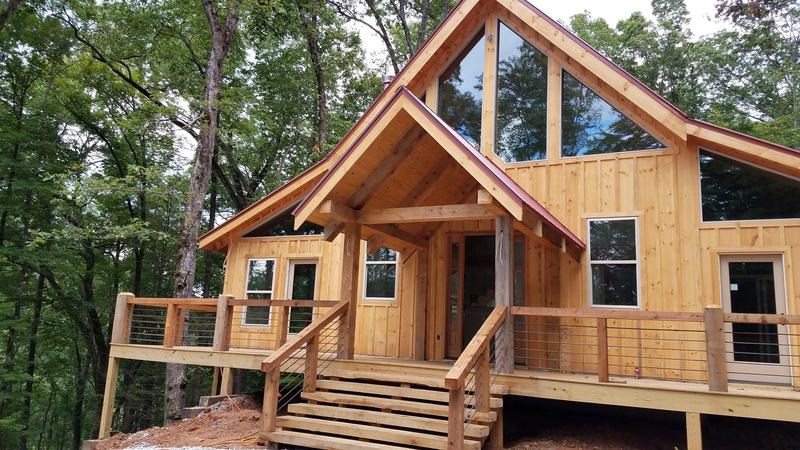 A new board-and-batten house in the woods with timber gables, tall windows and a wraparound porch