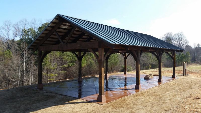 A dark-stained timber pavilion with curved braces and a black metal roof on a stained concrete pad. Opens larger.