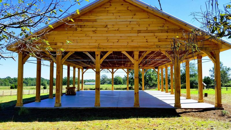 A long open pavilion with rows of timber posts and braces under a wood-plank gable. Opens larger.