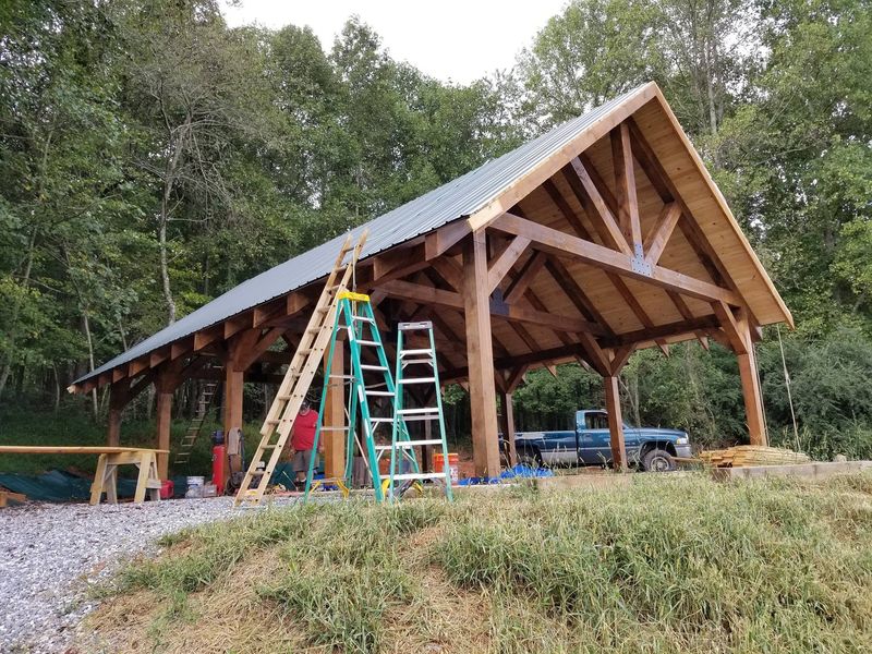 A timber pavilion frame with king-post trusses on a hillside, ladders set up under the roof. Opens larger.