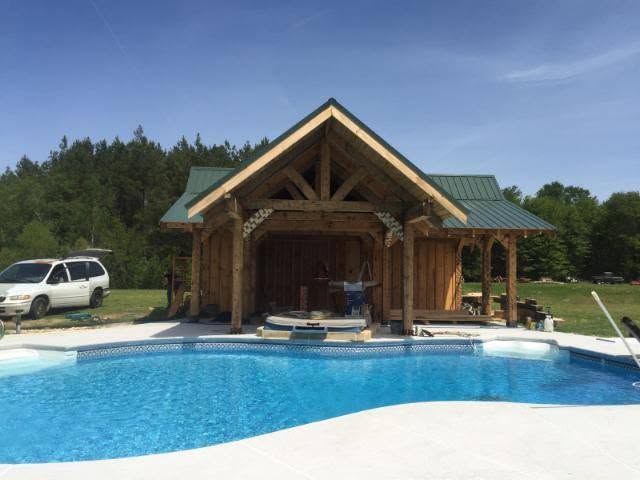 A timber-frame pool house with a green metal roof and an open gable, seen across a pool. Opens larger.