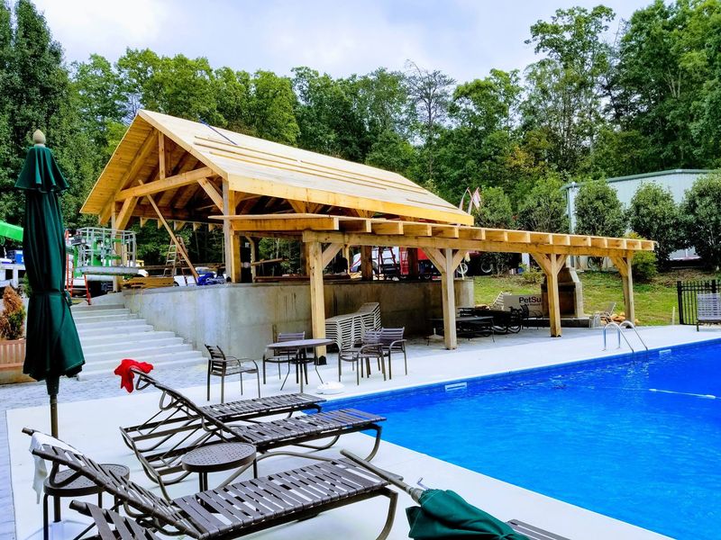 A large timber pavilion going up beside a pool, its frame and roof decking still bare. Opens larger.