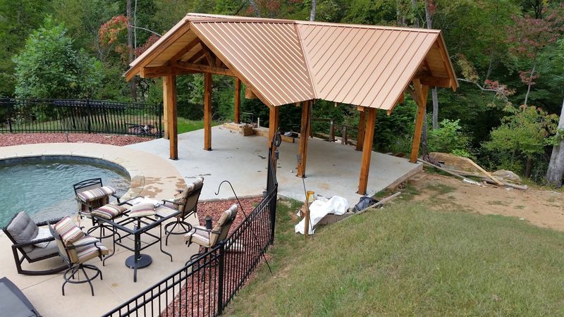 A timber pool pavilion with a copper-colored metal roof beside a pool and patio furniture. Opens larger.