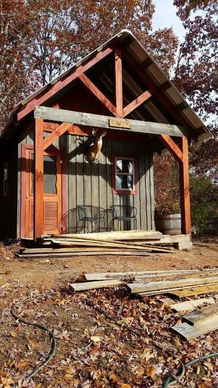 A small cabin with a red-stained timber gable porch, scrap lumber in the leaves in front. Opens larger.