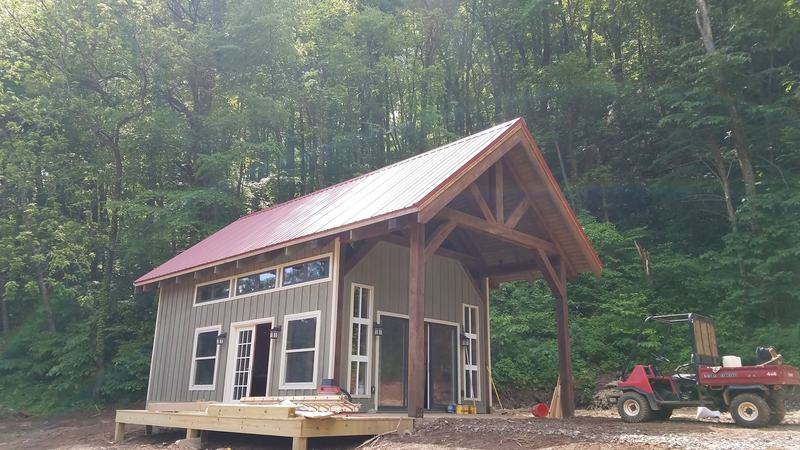 A small gray cabin with a red metal roof and a timber-frame porch gable. Opens larger.