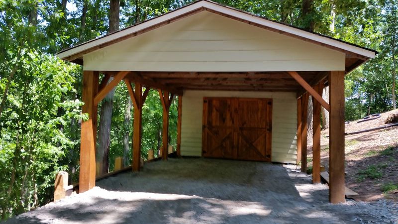 A timber-frame carport with heavy posts and curved braces, and a wood barn door on the back wall